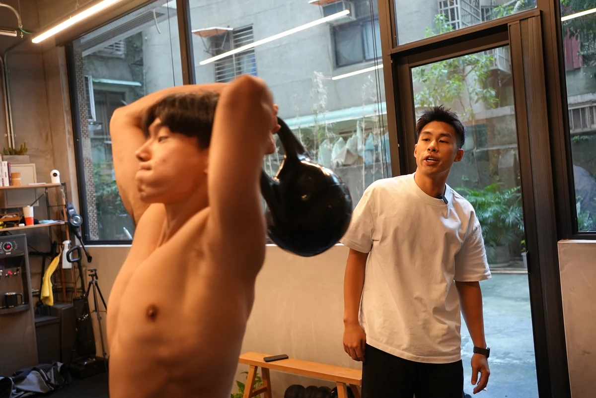 窗邊自然光下，學員做壺鈴過頭動作，Taka 在一旁看著
